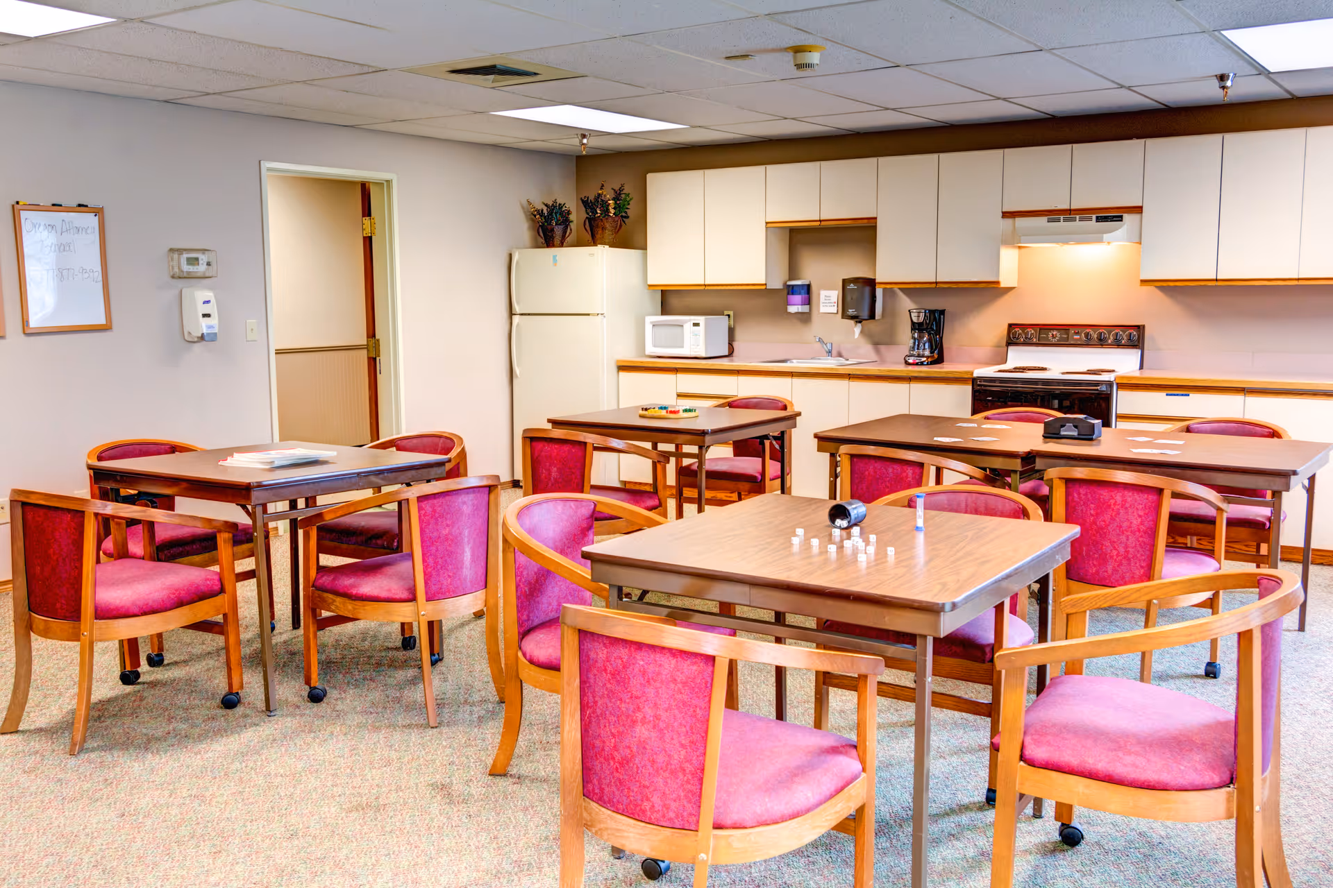 A communal room with several wooden tables and chairs with pink cushions arranged around them. The room includes a kitchenette area with white cabinets, a refrigerator, microwave, coffee maker, and stove. The walls are light-colored, and there is a whiteboard on one wall. The room appears well-lit and clean.