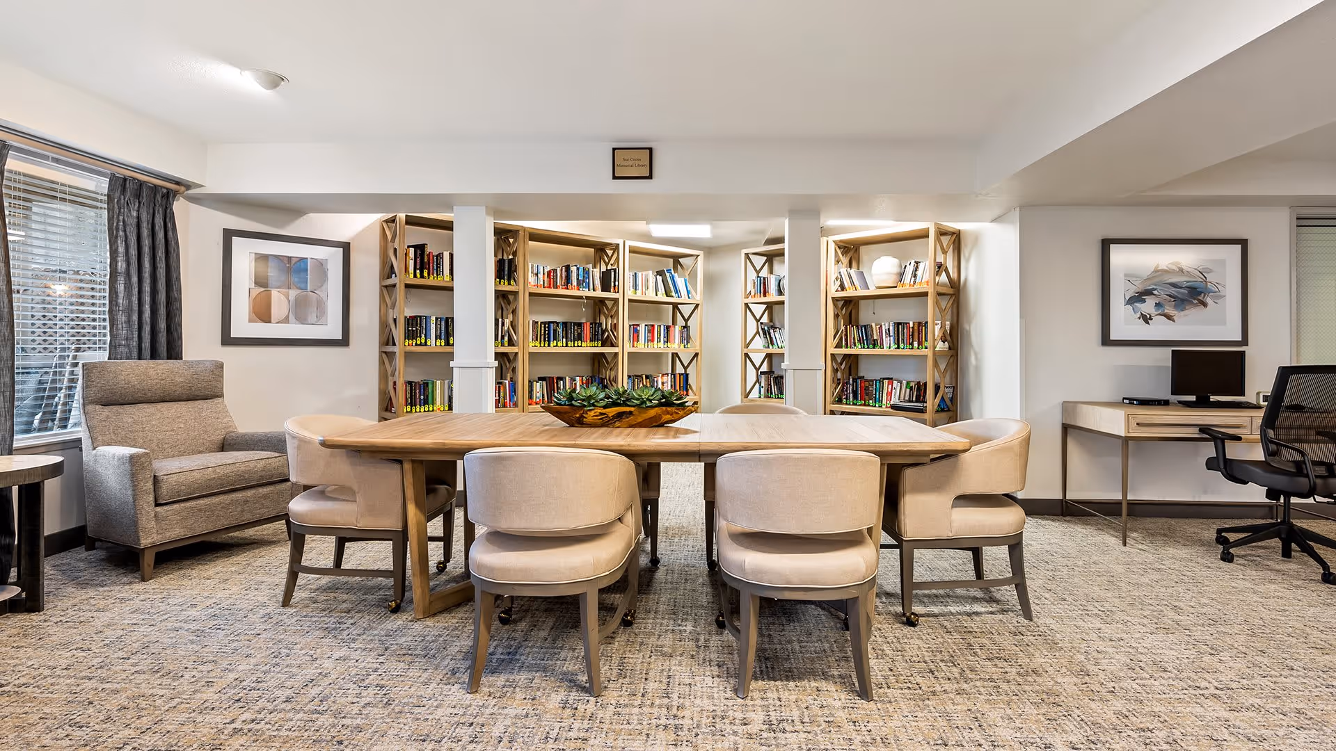 A cozy interior room with a large wooden table surrounded by eight beige upholstered chairs. Behind the table are multiple wooden bookshelves filled with books. To the left, there is a comfortable armchair next to a window with blinds and dark curtains. On the right side, there is a desk with a computer and an office chair. The walls are decorated with framed artwork.