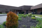 Exterior view of a single-story brick building with a flat roof, featuring a landscaped garden area with various shrubs and plants in the foreground under a clear sky.