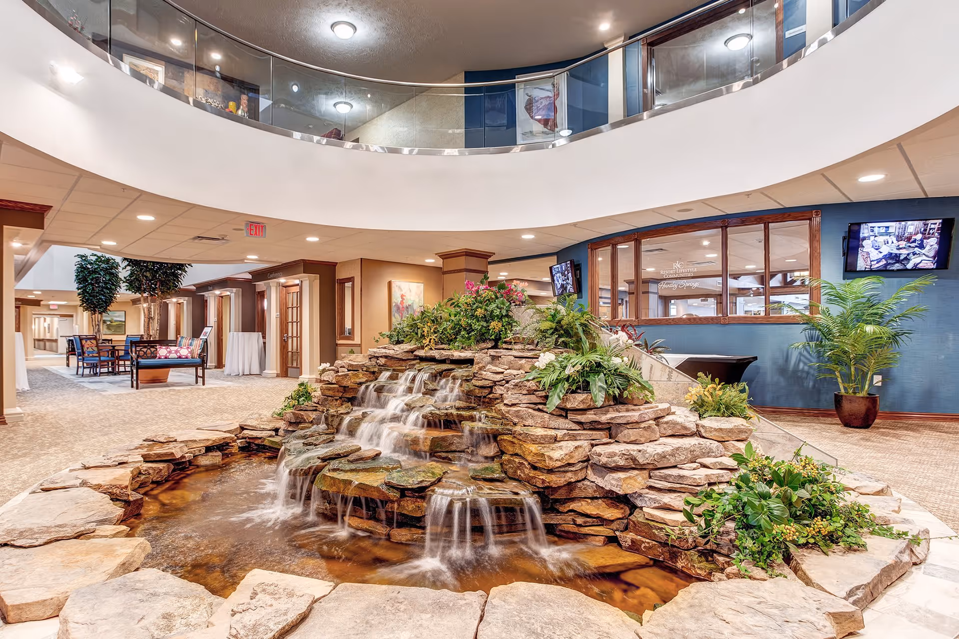 Indoor area of a senior living facility featuring a multi-tiered rock waterfall with flowing water surrounded by plants. The space has carpeted floors, seating areas with chairs and tables, and a second-floor balcony with glass railing overlooking the scene. Walls are decorated with artwork and there are two wall-mounted televisions.