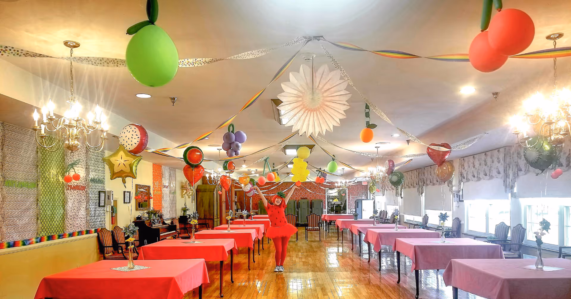 A large dining room decorated with colorful fruit-themed balloons and streamers hanging from the ceiling. The room has multiple tables covered with red tablecloths and chairs arranged around them. A person dressed in a red costume stands in the center of the room with arms raised. The room is well-lit with chandeliers and natural light from windows on the right side.