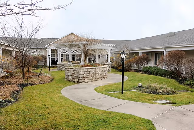 Outdoor courtyard area at a senior living facility with a curved concrete walkway, green grass, leafless trees, shrubs, a stone planter, a white pergola, and a building with a gray roof in the background under an overcast sky.