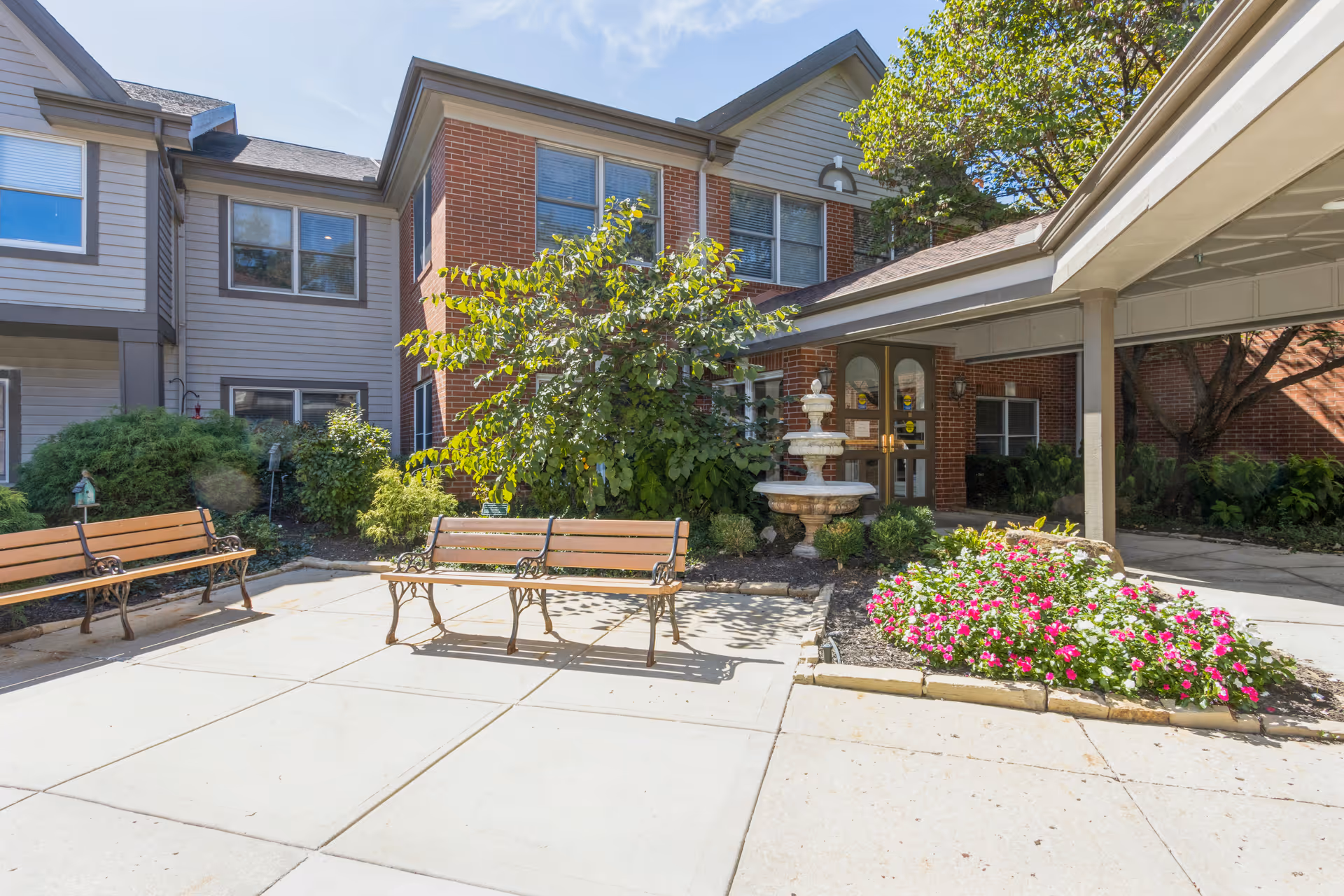 Outdoor courtyard area of a senior living facility with two wooden benches, a stone fountain, flower beds with pink and white flowers, and a covered entrance to a brick and siding building under a clear blue sky.