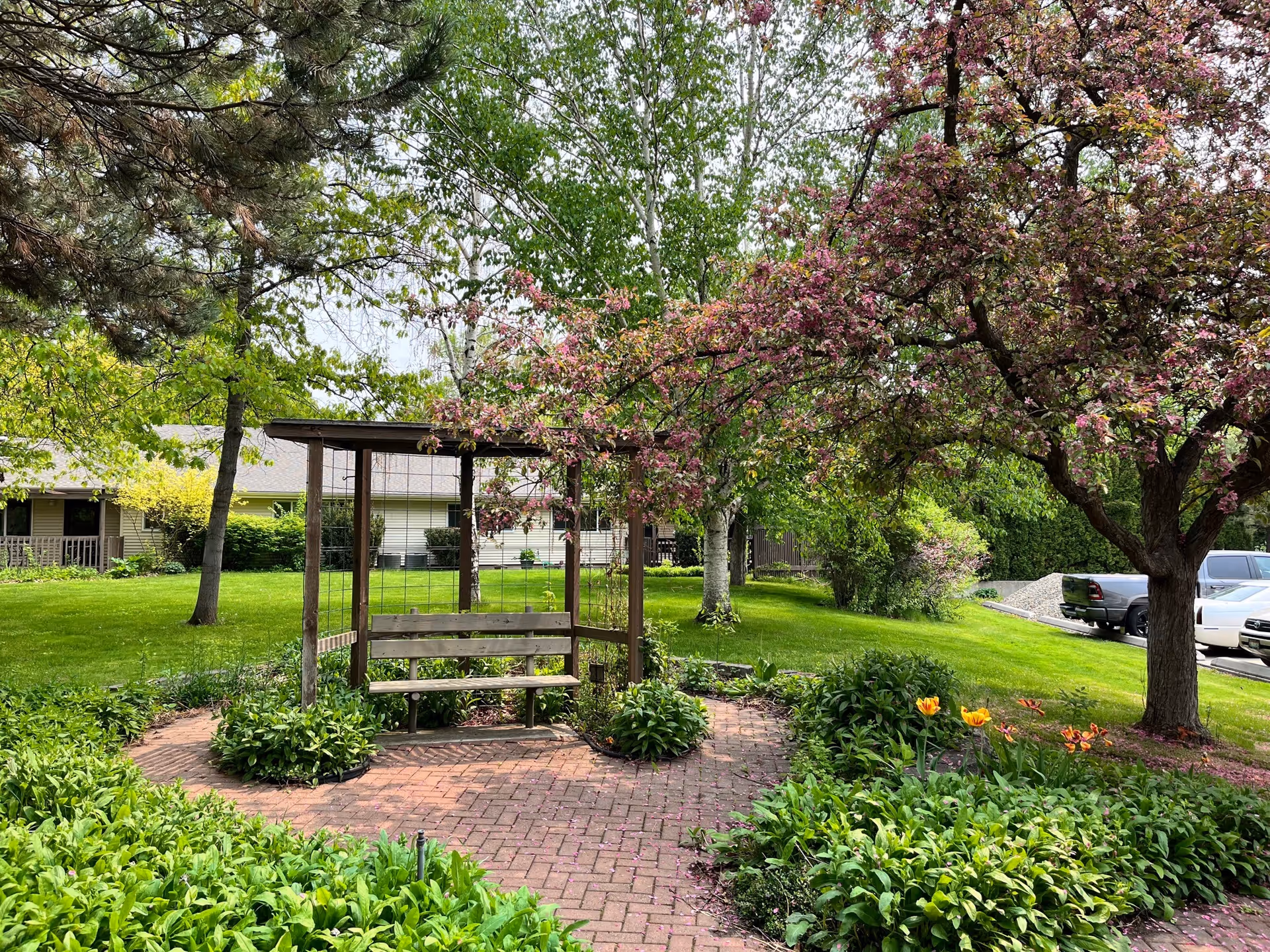 Brick-paved garden with a wooden arbor bench surrounded by flowering trees, shrubs, and lawn in front of a retirement building.