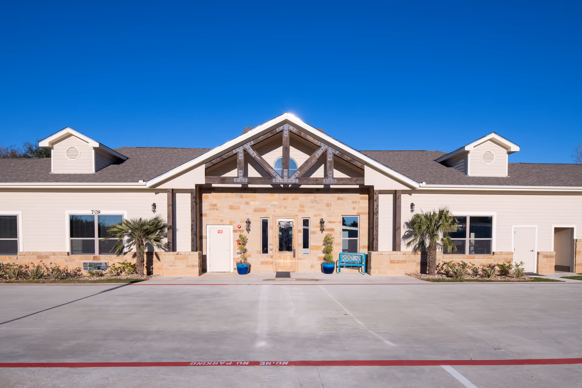 Front exterior view of a single-story assisted living facility building with a peaked roof, beige siding, stone accents around the entrance, two small palm trees, and a blue bench near the entrance under a clear blue sky.
