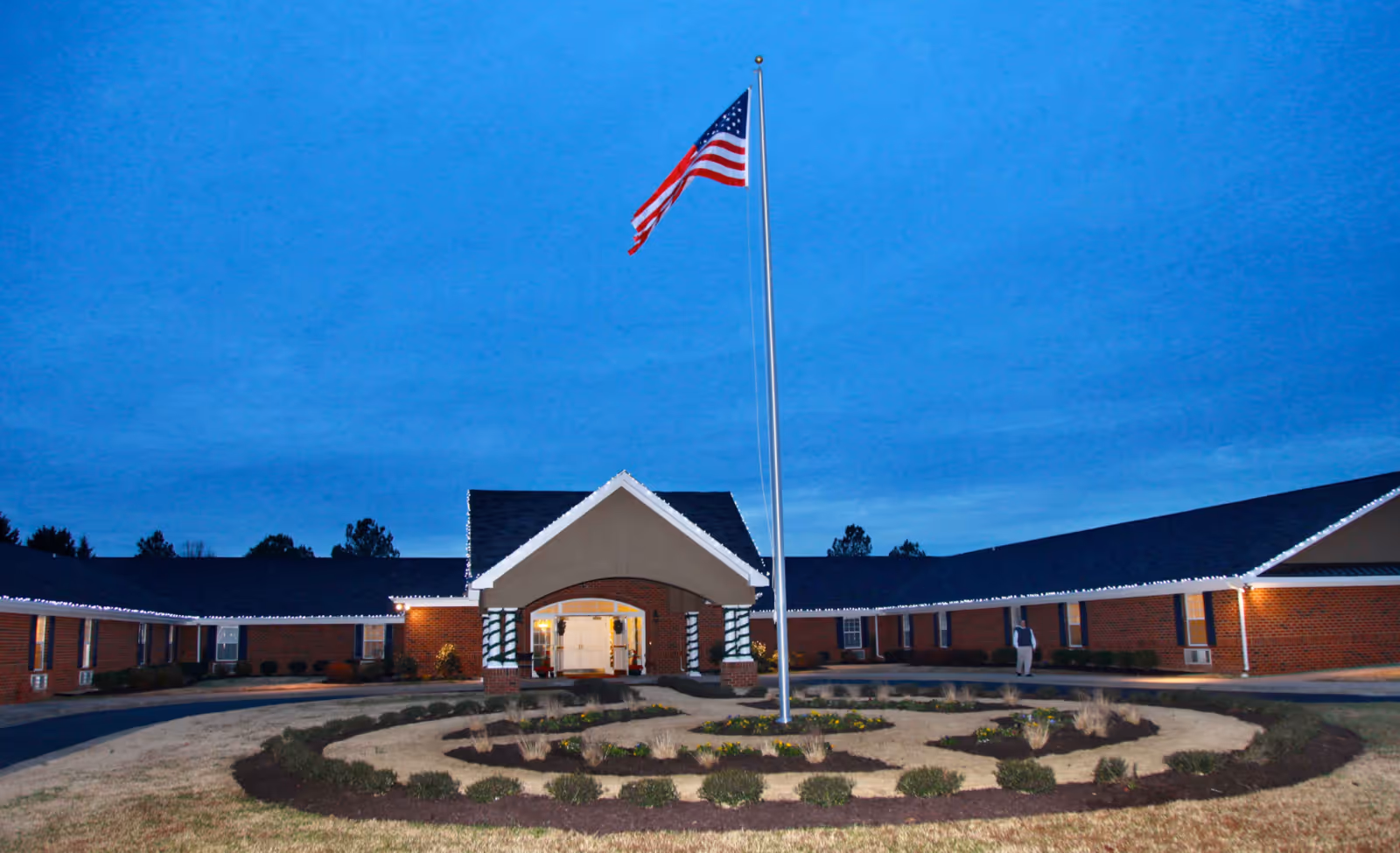 Exterior view of Commonwealth Senior Living at Chesterfield building at dusk with an American flag on a tall flagpole in the center of a circular landscaped area in front of the entrance. The building is a single-story brick structure with a dark roof and white trim, decorated with white string lights along the roofline.