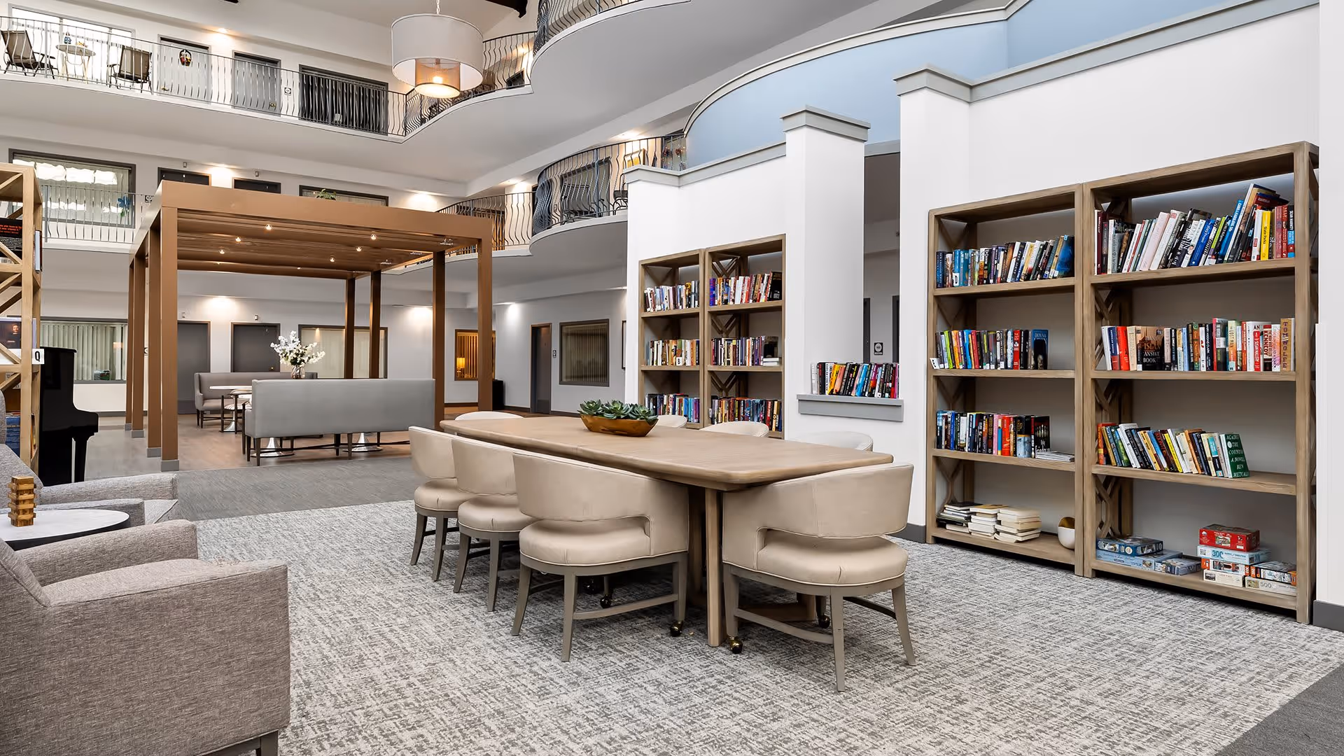 A spacious and well-lit common area in a senior living facility featuring a long wooden table with beige chairs, bookshelves filled with books and board games, comfortable armchairs, and a wooden pergola structure with additional seating in the background. The area has a high ceiling with a second-floor balcony overlooking the space.