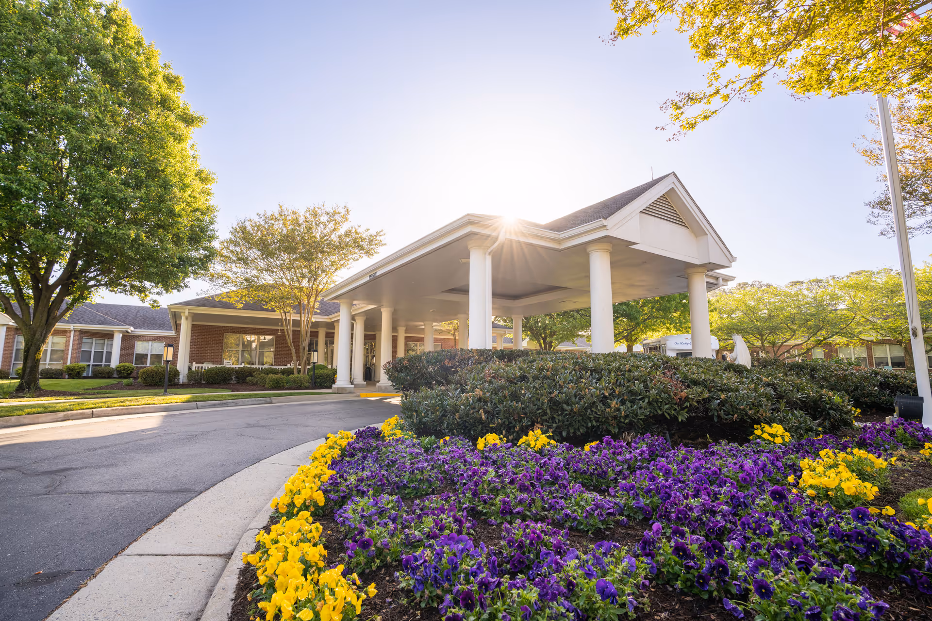 Entrance of a senior living facility with a covered drop-off area supported by white columns. The foreground features a vibrant flower bed with purple and yellow flowers, and there are green trees and shrubs surrounding the building. The sun is shining brightly in the clear sky behind the building.