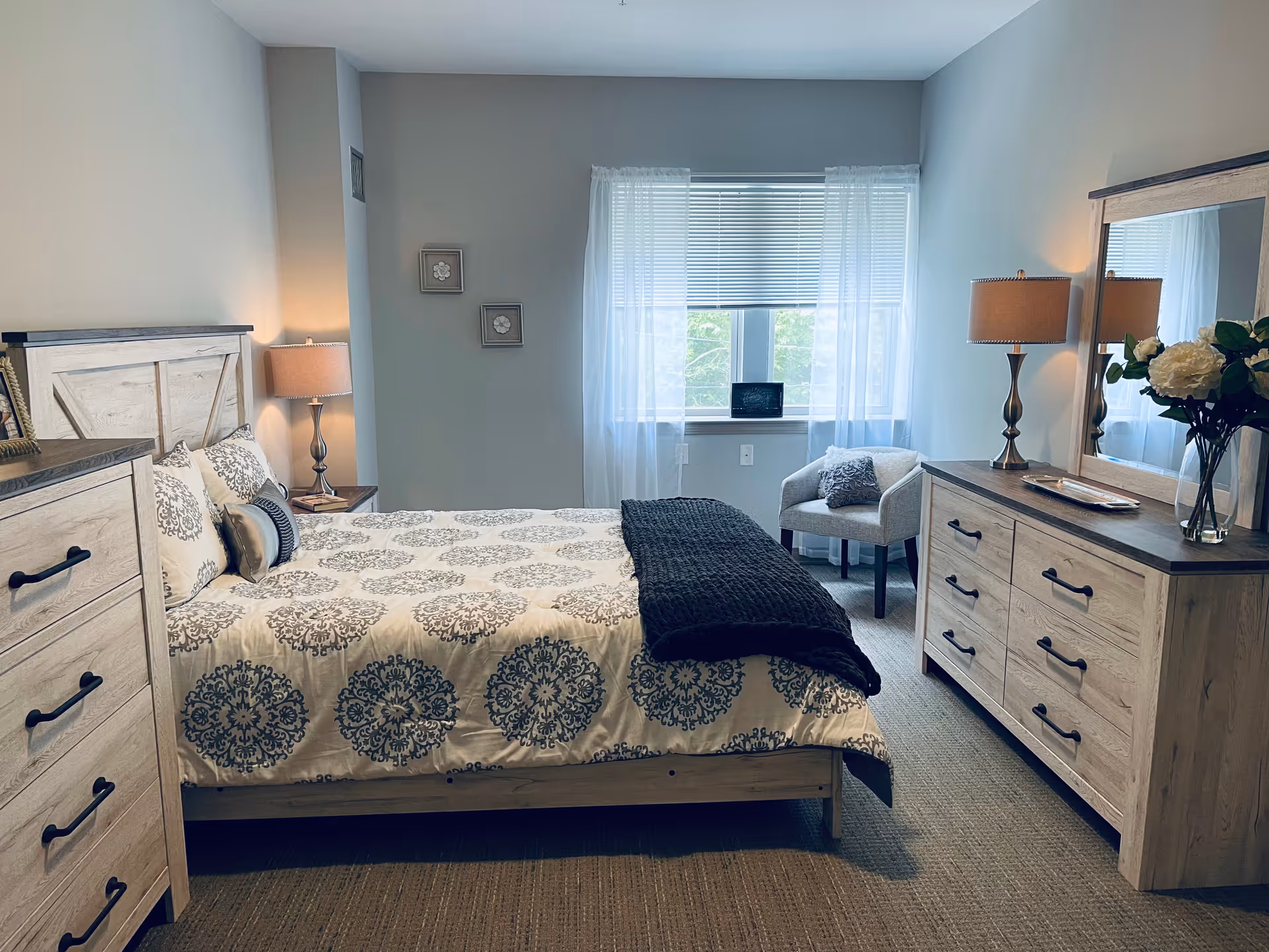 A neatly arranged bedroom with a bed featuring a patterned white and gray bedspread and a dark throw blanket. The room includes a wooden dresser with a mirror, a matching chest of drawers, two table lamps, a small armchair near a window with sheer white curtains and blinds, and two framed pictures on the wall.