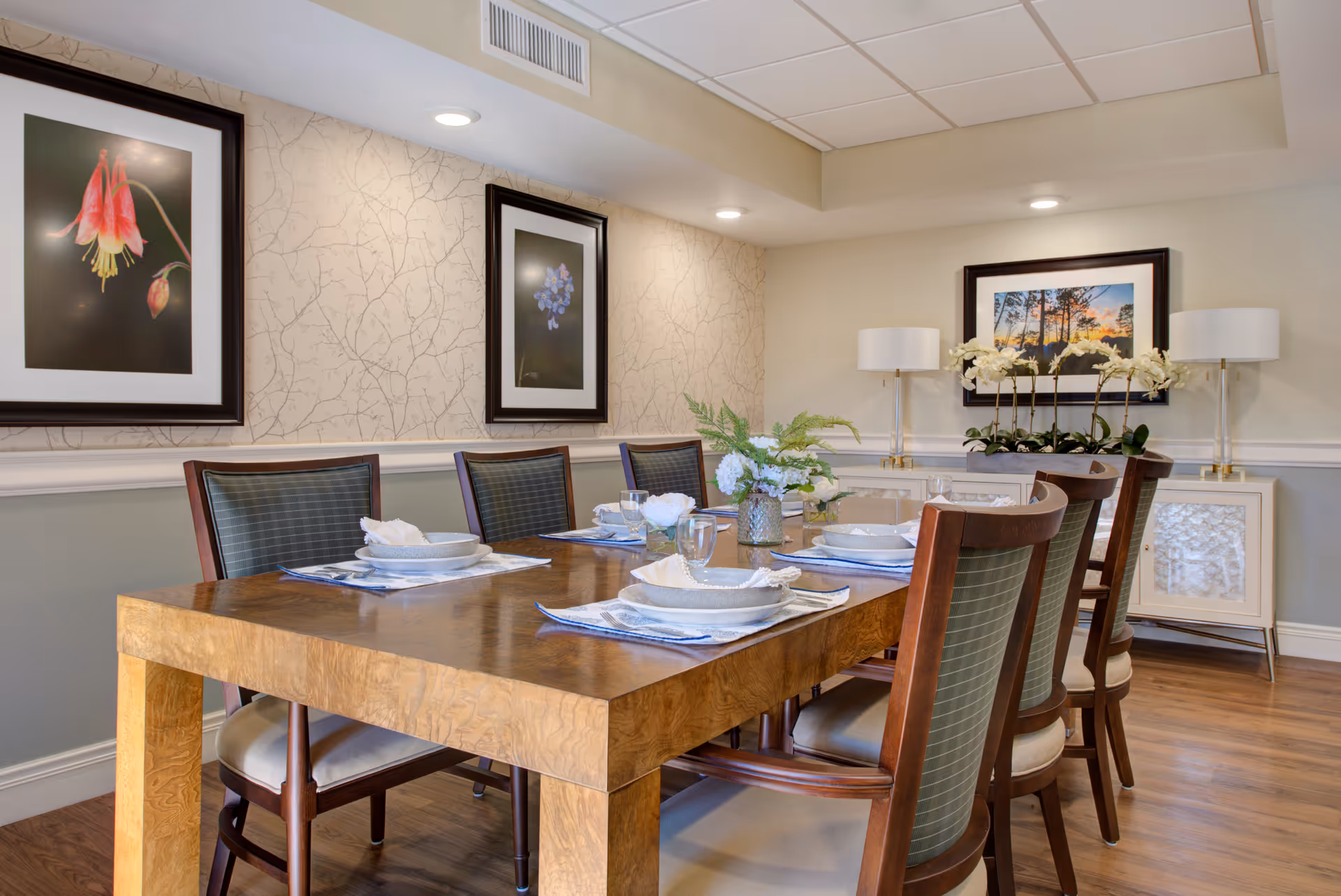 A dining room with a wooden table set for six people, featuring plates, bowls, glasses, and napkins. The room has six chairs with cushioned seats and backs, floral artwork on the walls, two table lamps on a sideboard, and decorative plants on the table and sideboard. The floor is wooden and the walls are painted in neutral tones with patterned wallpaper on one side.