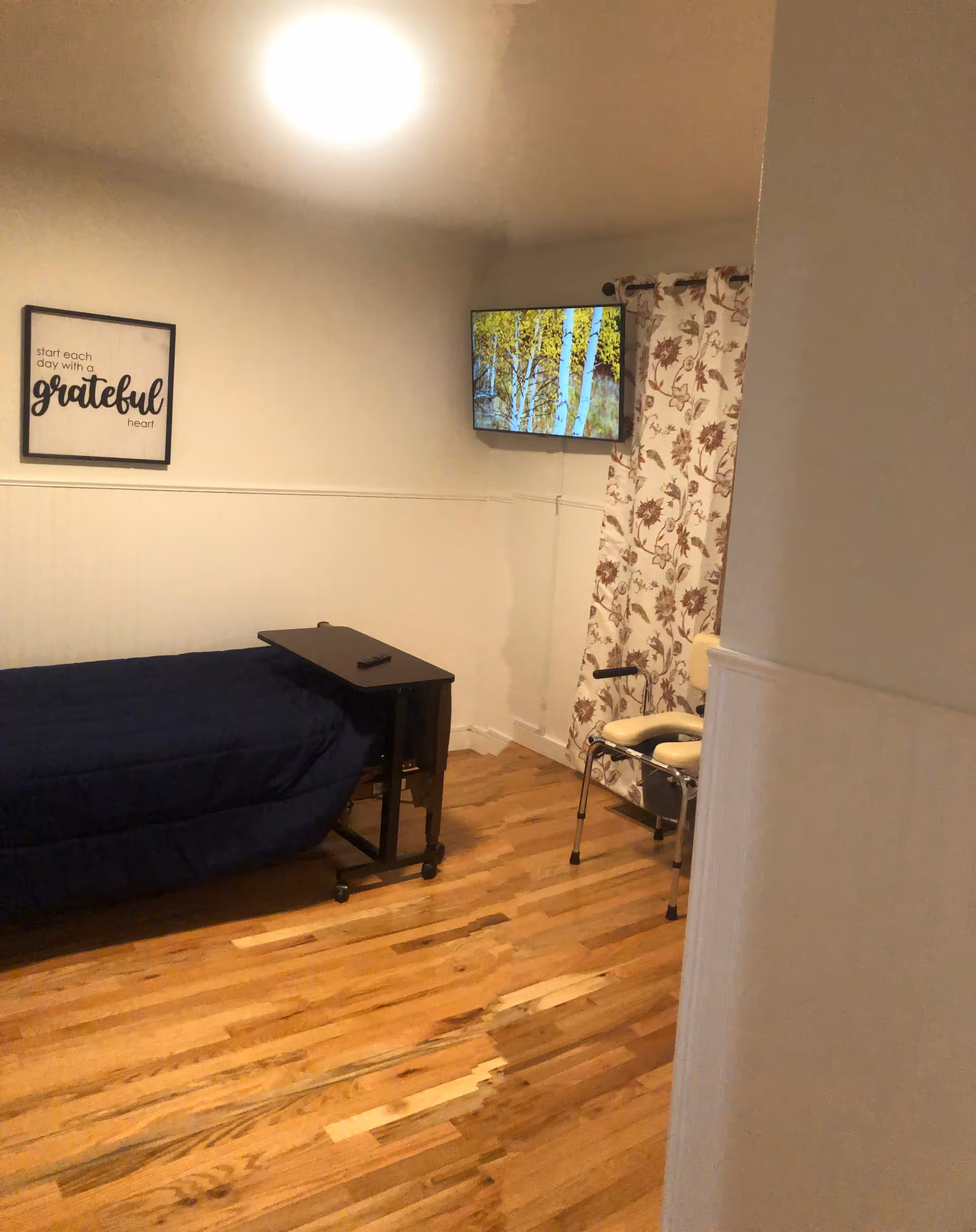 A small bedroom with a single bed covered in a dark blue comforter, a black rolling overbed table, a wall-mounted TV displaying an image of birch trees, a chair with armrests, floral curtains, and a framed wall art that reads 'start each day with a grateful heart'. The floor is wooden and the walls are painted white.