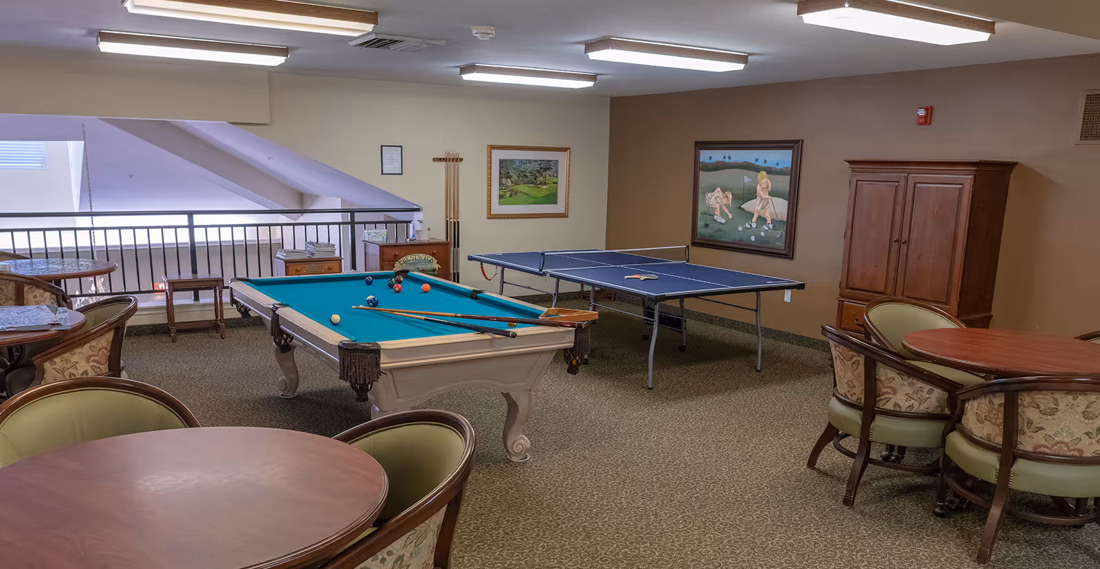 A recreational room in a senior living community featuring a pool table with cues and balls, a ping pong table with paddles, several round tables with cushioned chairs, framed artwork on the walls, and a wooden cabinet. The room has carpeted flooring and fluorescent ceiling lights.