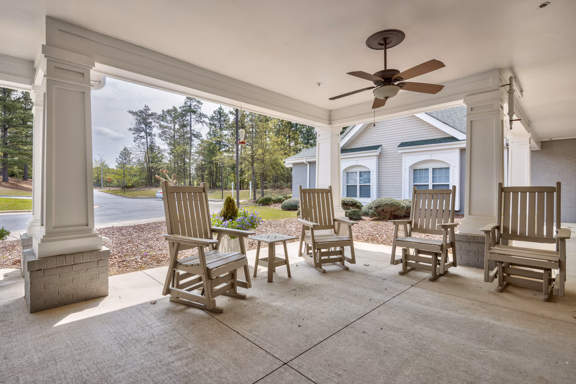 Covered outdoor seating area with four wooden rocking chairs and two small tables, ceiling fan above, surrounded by white pillars. In the background, there are trees, shrubs, and a building with white siding and green roof.