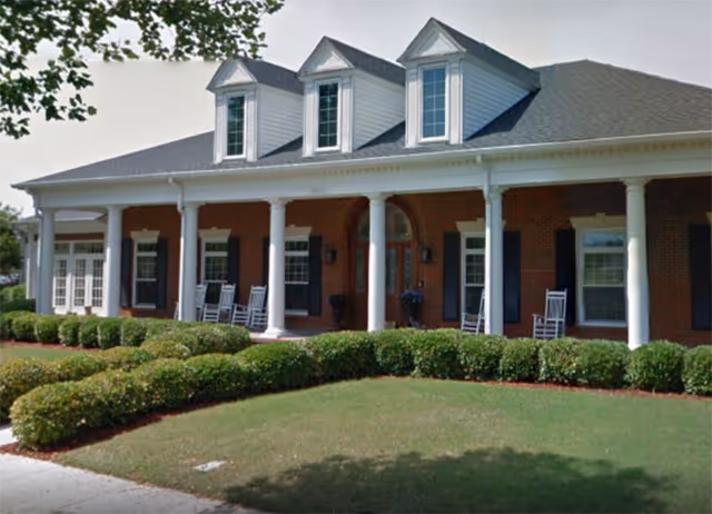 Front exterior view of a traditional brick building with white columns and a covered porch featuring several white rocking chairs. The building has three dormer windows on the roof and is surrounded by neatly trimmed bushes and a green lawn.