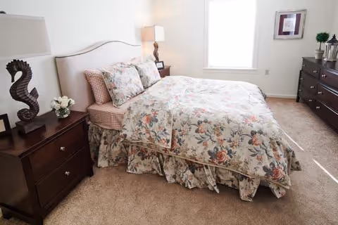 A bedroom with a bed covered in floral patterned bedding and pillows. There are two dark wooden nightstands on either side of the bed, each with a lamp. One nightstand has a decorative seahorse sculpture and a small vase of white flowers. A window with white blinds is on the wall behind the bed, and a dark wooden dresser with a plant and framed picture is on the right side of the room.