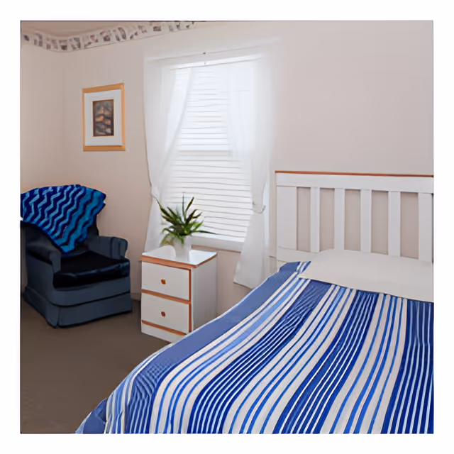 A cozy bedroom featuring a bed with a blue and white striped bedspread, a white headboard, a small white nightstand with two drawers and a potted plant on top, a window with white curtains, and a blue armchair with a blue patterned throw draped over it. A framed picture hangs on the wall above the armchair.