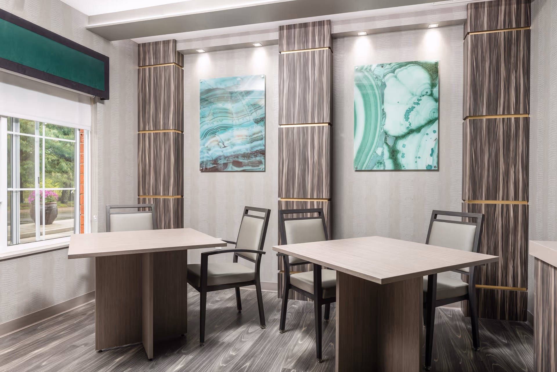 A modern dining area with two square wooden tables and four chairs with light-colored cushions. The room features large windows with a green valance, wood-patterned vertical wall panels with gold accents, and two abstract green and white artworks on the wall. The floor has a gray wood-like finish.
