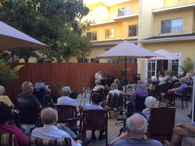 Residents seated in a courtyard watching a musician perform under umbrellas outside a senior living building.