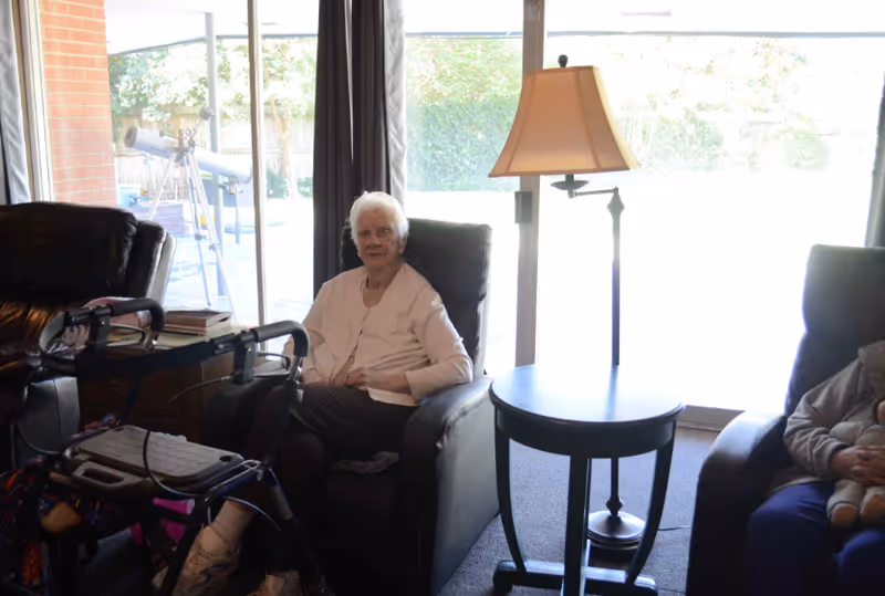An elderly woman seated in a recliner in a sunlit common living area with a side table, lamp, and a walker nearby.