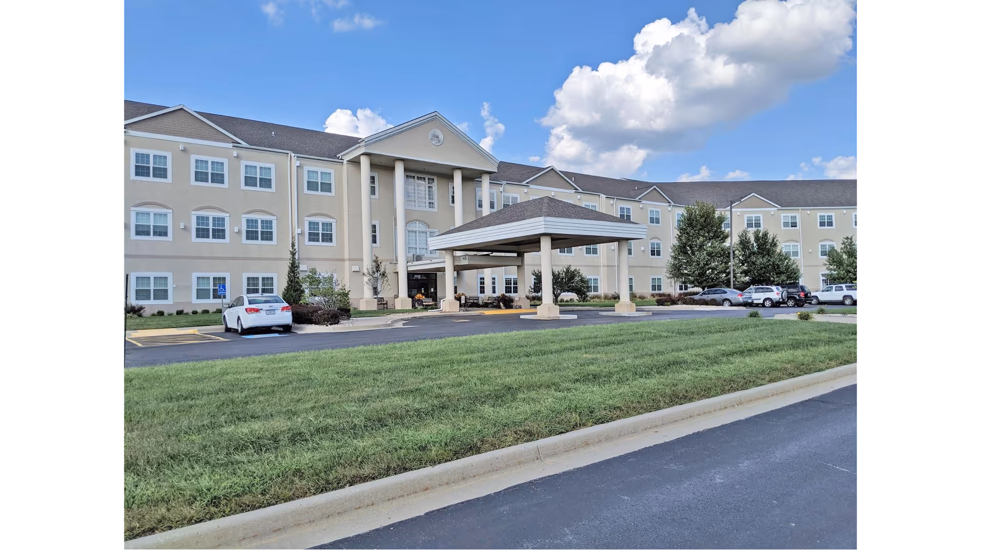 Exterior view of a three-story beige retirement community building with multiple windows, a covered entrance with columns, a parking lot with several cars, green grass in the foreground, and a partly cloudy blue sky.