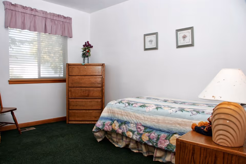 A small bedroom with a single bed covered in floral bedding, a wooden dresser with a vase of flowers on top, a wooden chair, a window with blinds and a pink valance, two framed pictures on the wall, and a bedside table with a lamp.