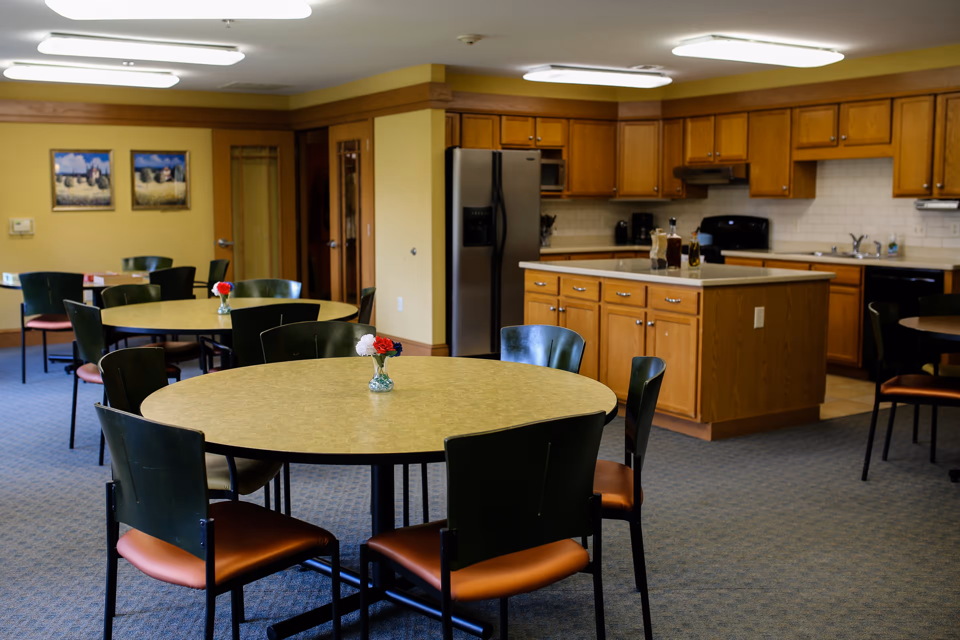A communal dining area with round tables and chairs, each table decorated with a small vase containing red and white flowers. In the background, there is a kitchen with wooden cabinets, a refrigerator, stove, and a kitchen island. The walls are painted yellow and there are two framed pictures hanging on the wall.