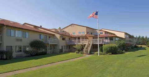 Two-story senior living facility exterior with balconies, green lawn, and an American flag.
