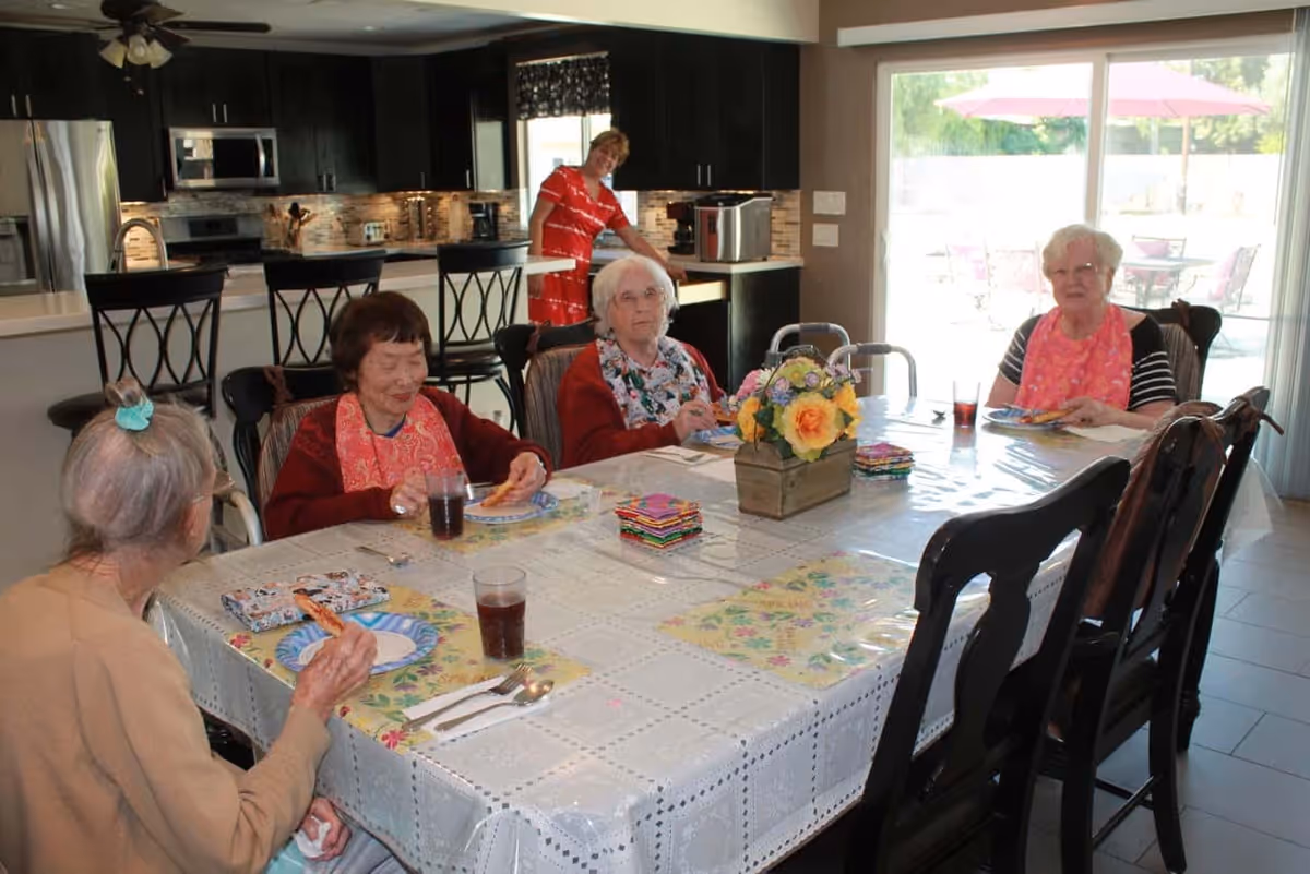 Four elderly women sitting around a dining table with floral placemats and a flower centerpiece, eating pizza and drinking beverages. A woman in a red dress stands in the kitchen area behind them. The room has a modern kitchen with dark cabinets and stainless steel appliances, and a sliding glass door leading to an outdoor patio with tables and umbrellas.