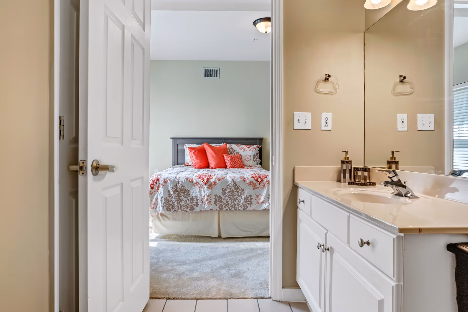 View through an open door showing a bedroom with a bed dressed in a patterned comforter and red pillows. To the right is a bathroom vanity with a sink, soap dispenser, and a large mirror reflecting part of the room.