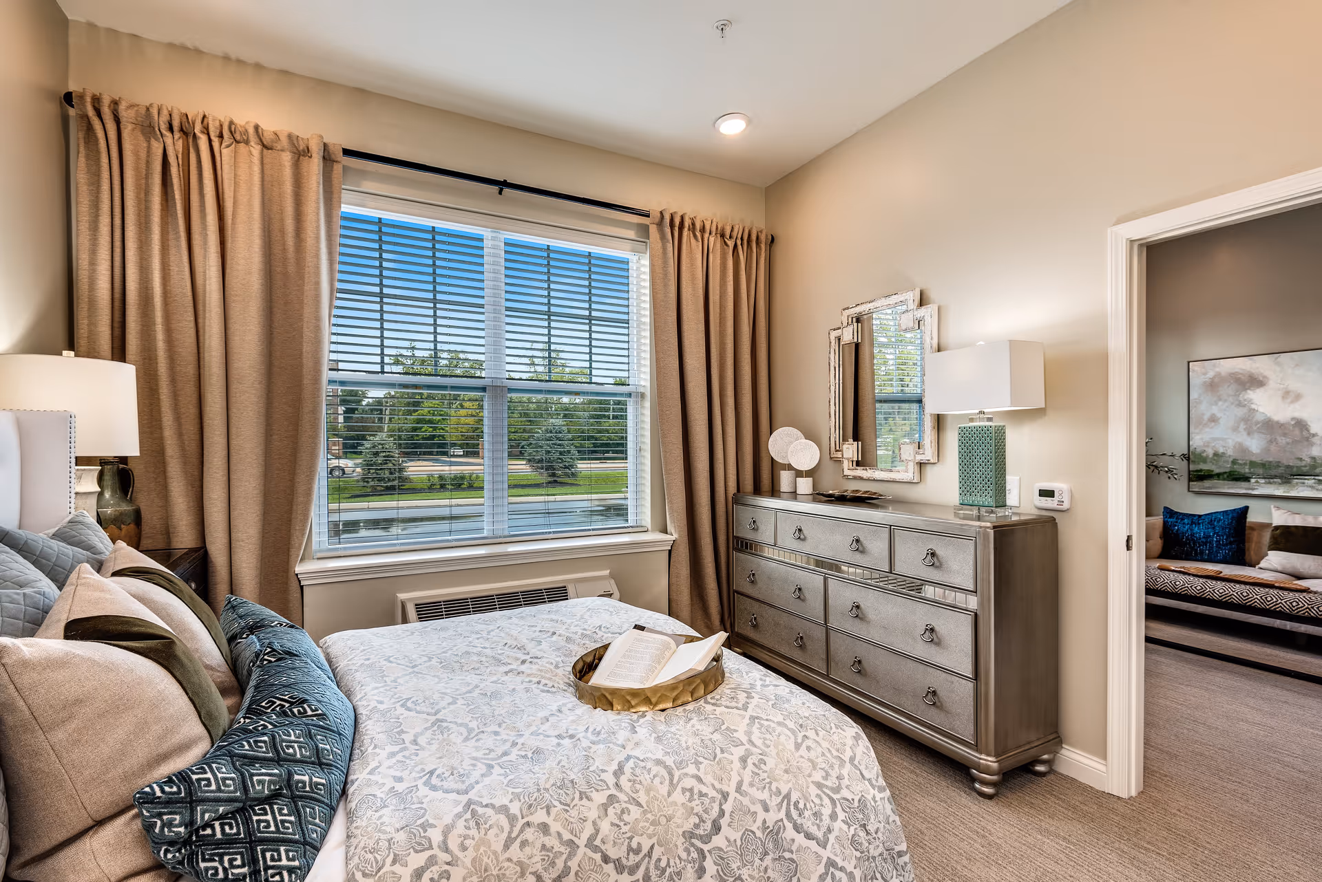 A cozy bedroom with a large window covered by beige curtains and blinds. The bed is neatly made with patterned bedding and multiple pillows, and a tray with an open book rests on the bed. A metallic dresser with a decorative mirror and a lamp is positioned against the wall. An open doorway leads to another room with a sofa and artwork on the wall.
