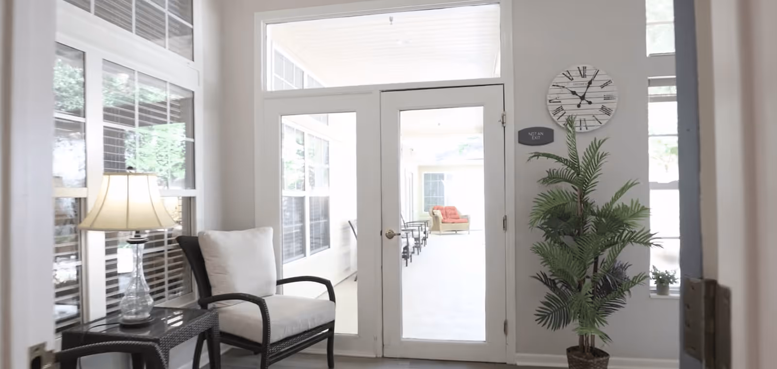 A bright interior room featuring a cushioned armchair next to a small table with a lamp, a large potted plant, and glass double doors leading to a sunlit seating area with more chairs and a sofa with orange cushions.