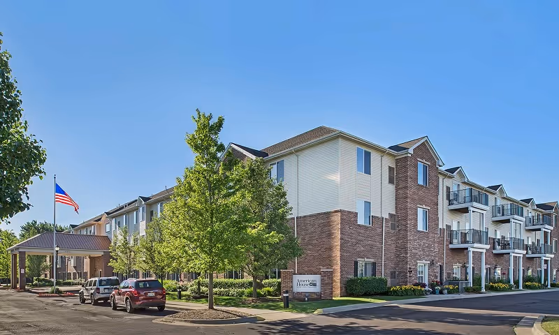 Three-story brick-and-siding senior living building with balconies, a landscaped entrance, parked cars, and an American flag under a clear blue sky.