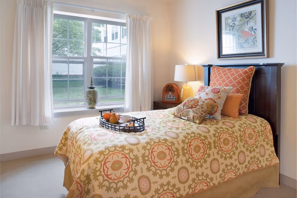 A cozy bedroom with a single bed covered in a patterned bedspread with multiple decorative pillows. A tray with fruit and a cup is placed on the bed. There is a window with white curtains letting in natural light, a bedside table with a lamp and a vintage radio, and a framed artwork hanging on the wall above the bed.