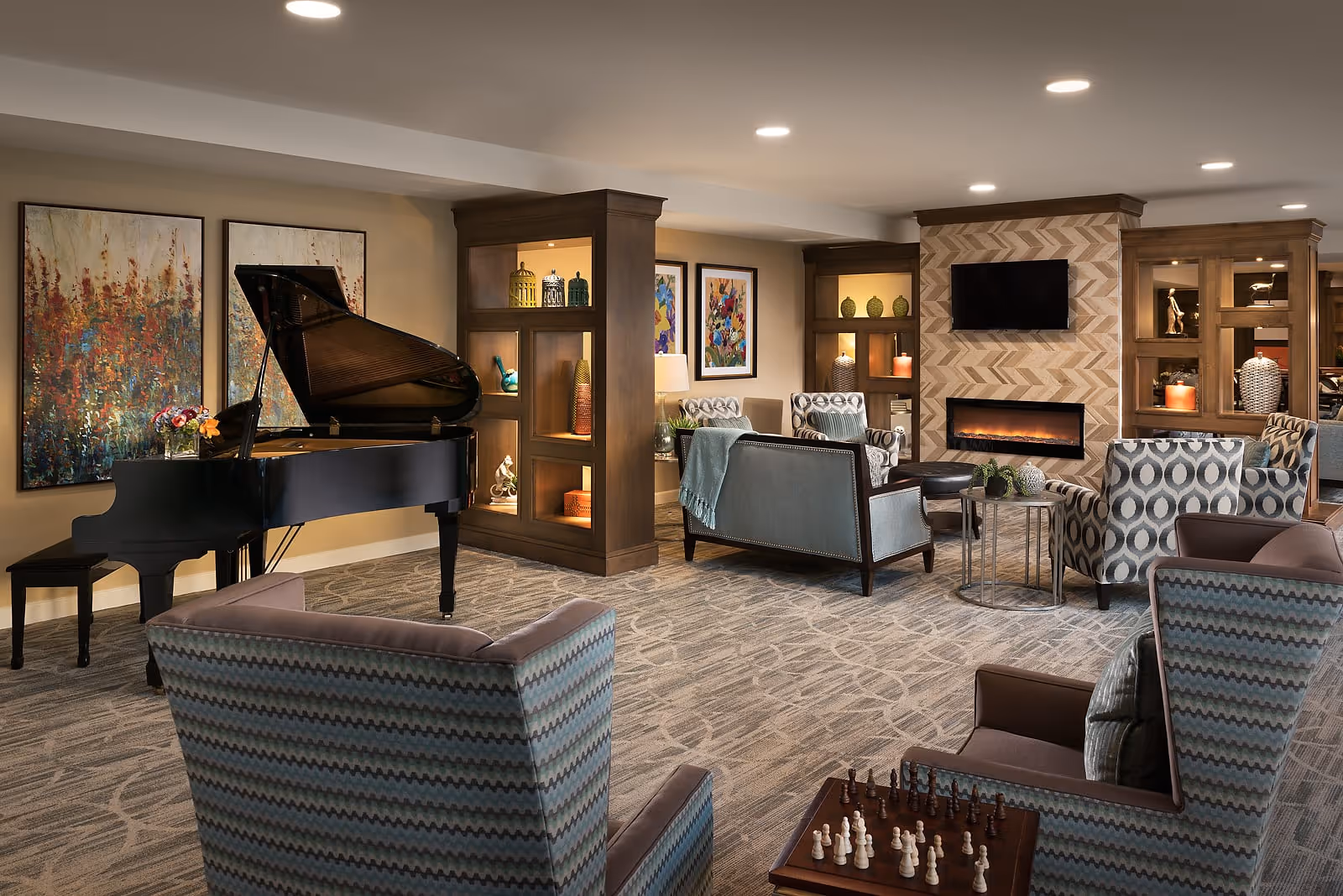 A spacious and elegantly decorated living room with a grand piano on the left side, several comfortable armchairs and sofas arranged around a modern electric fireplace with a TV mounted above it. The room features built-in wooden shelves with decorative items and colorful artwork on the walls. A chessboard is set up on a small table in the foreground.
