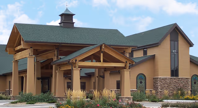 Exterior view of a senior living facility building with a large covered entrance supported by wooden beams, beige walls, stone accents, and a green roof under a partly cloudy sky.