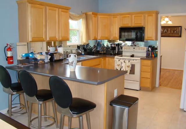 A kitchen with light wood cabinets, a gray countertop island with three black bar stools, a white stove with butterfly-themed towels, a microwave above the stove, a window with a beige curtain, various kitchen appliances and utensils on the counters, a fire extinguisher mounted on the wall, and a trash can next to the island.