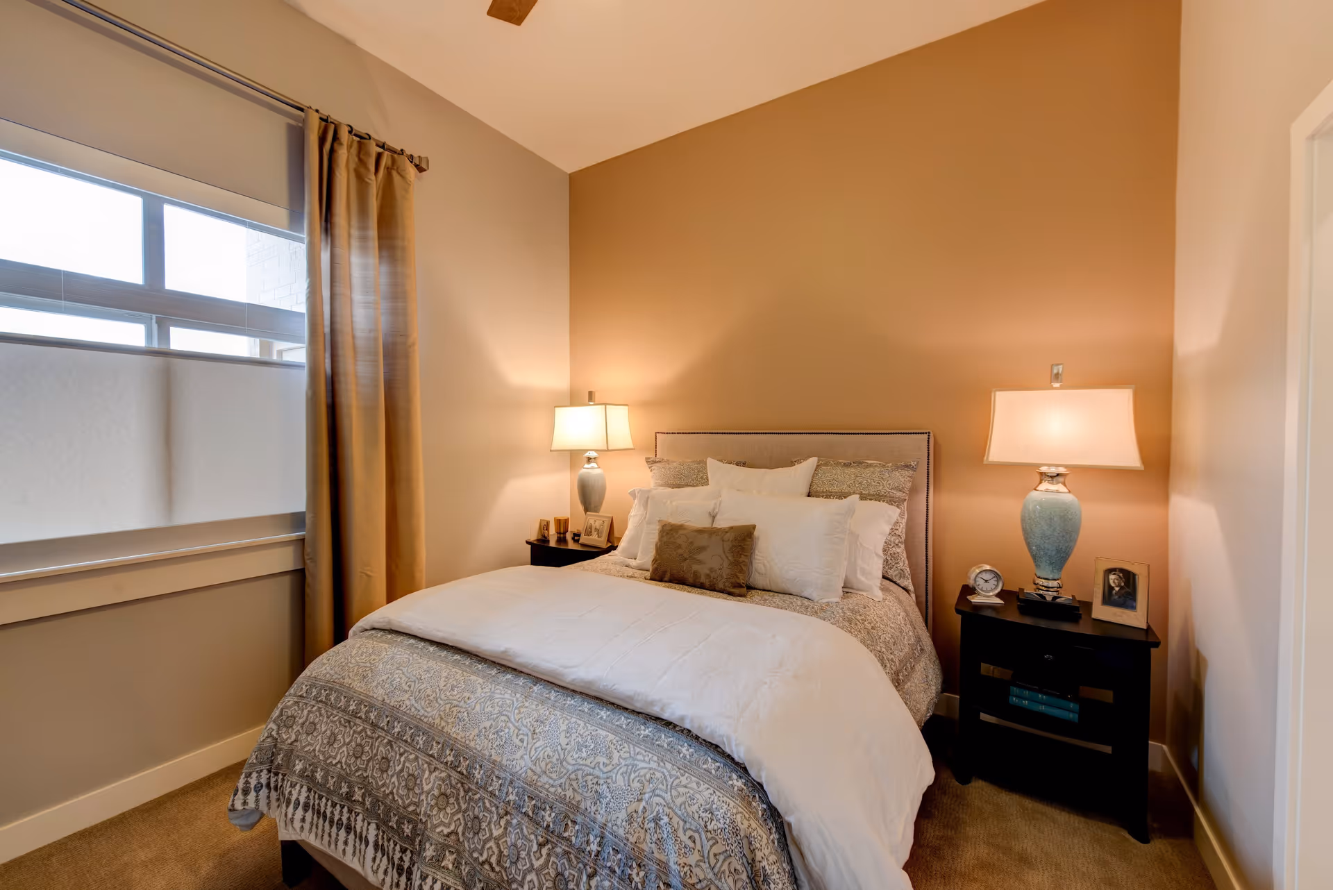 A cozy bedroom with a neatly made bed featuring patterned bedding and multiple pillows. There are two bedside tables, each with a lamp, a small clock, and framed photos. A window with a curtain and a roller shade is on the left wall, and the walls are painted in warm neutral tones.