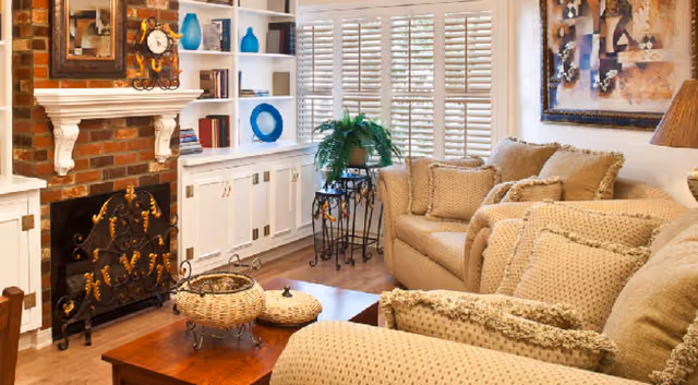 Cozy living room with beige upholstered sofas, a brick fireplace with white mantel, built-in shelving, coffee table, and shuttered window.
