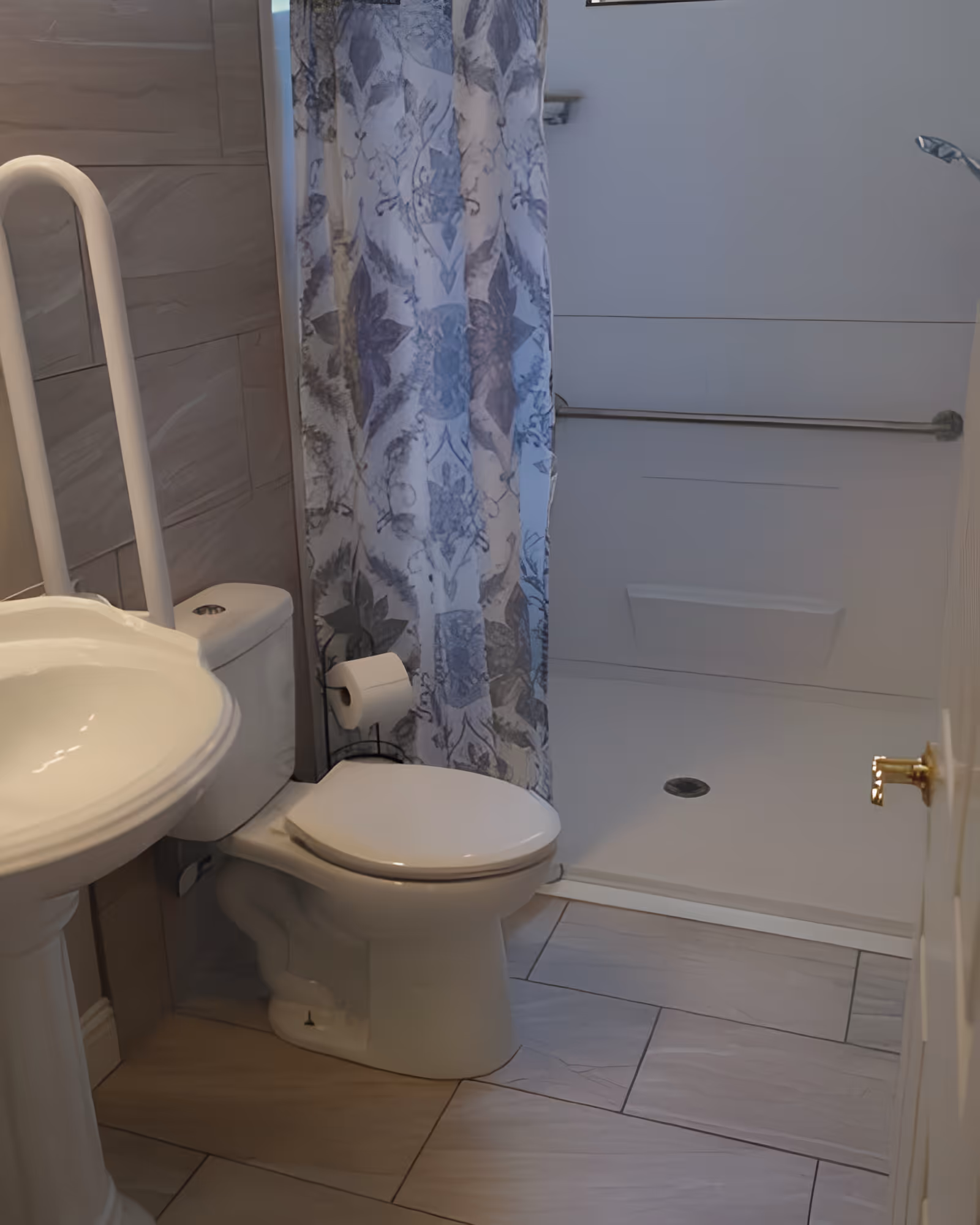A bathroom with a white pedestal sink, a toilet with a grab bar beside it, a toilet paper holder, and a walk-in shower with a floral patterned shower curtain and a horizontal grab bar inside the shower.