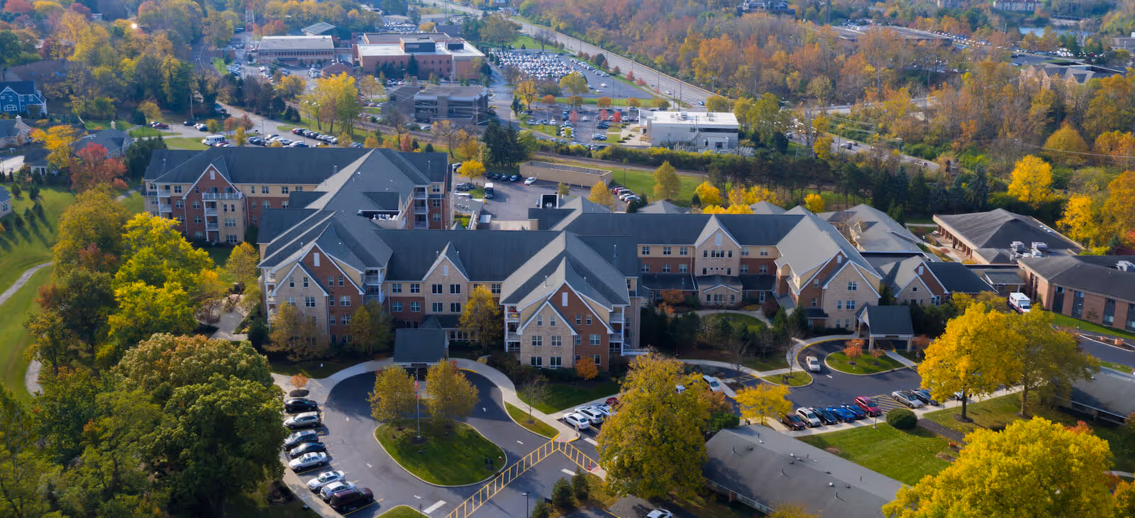 Aerial view of a large senior living facility named First Community Village surrounded by trees with autumn foliage. The facility consists of multiple connected buildings with gray roofs and brick facades. There are parking lots with cars, landscaped green areas, and roads around the property. In the background, there are more buildings and a highway.