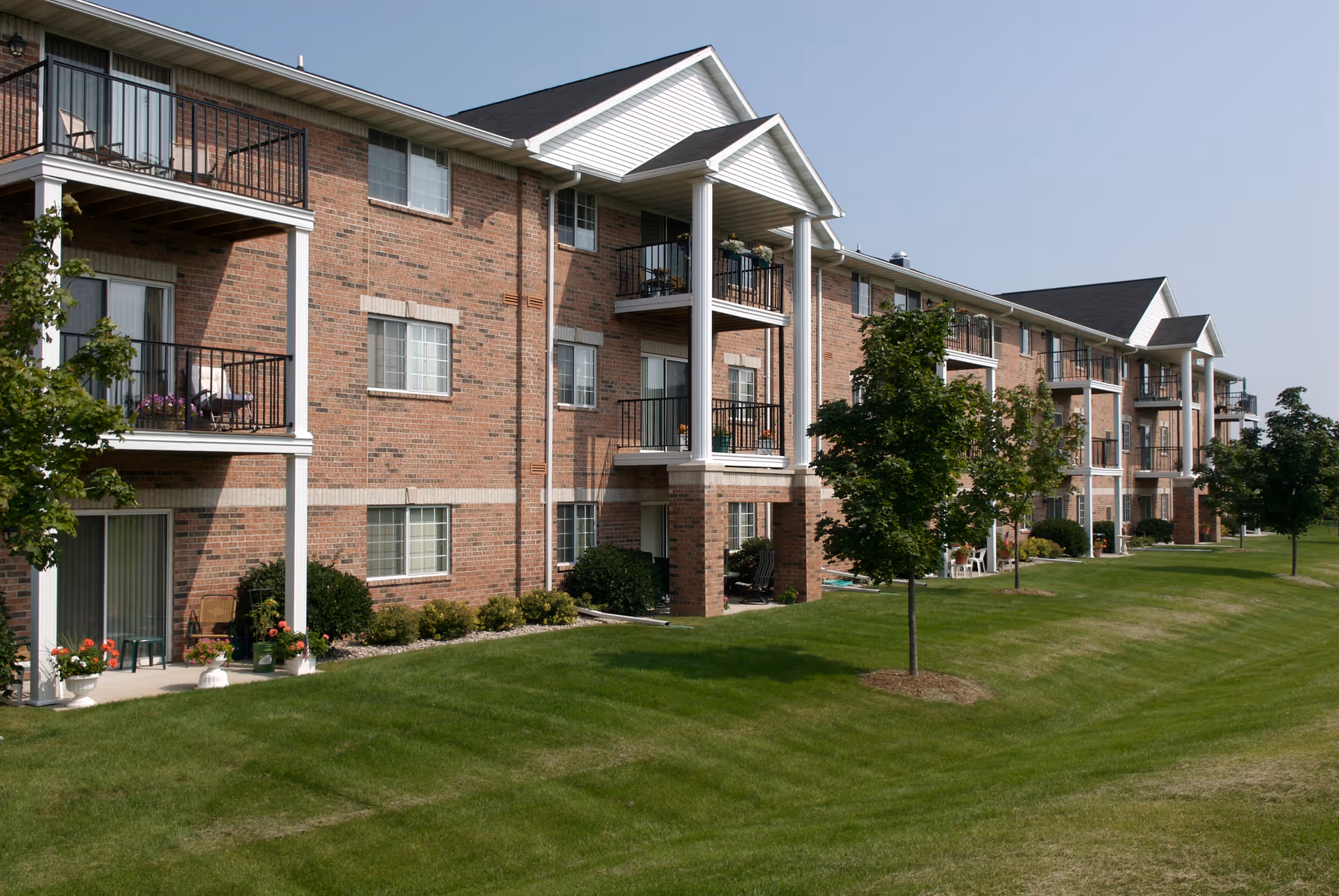 Exterior view of a three-story brick apartment building with balconies and patios. The building is surrounded by a well-maintained lawn with small trees and shrubs under a clear blue sky.