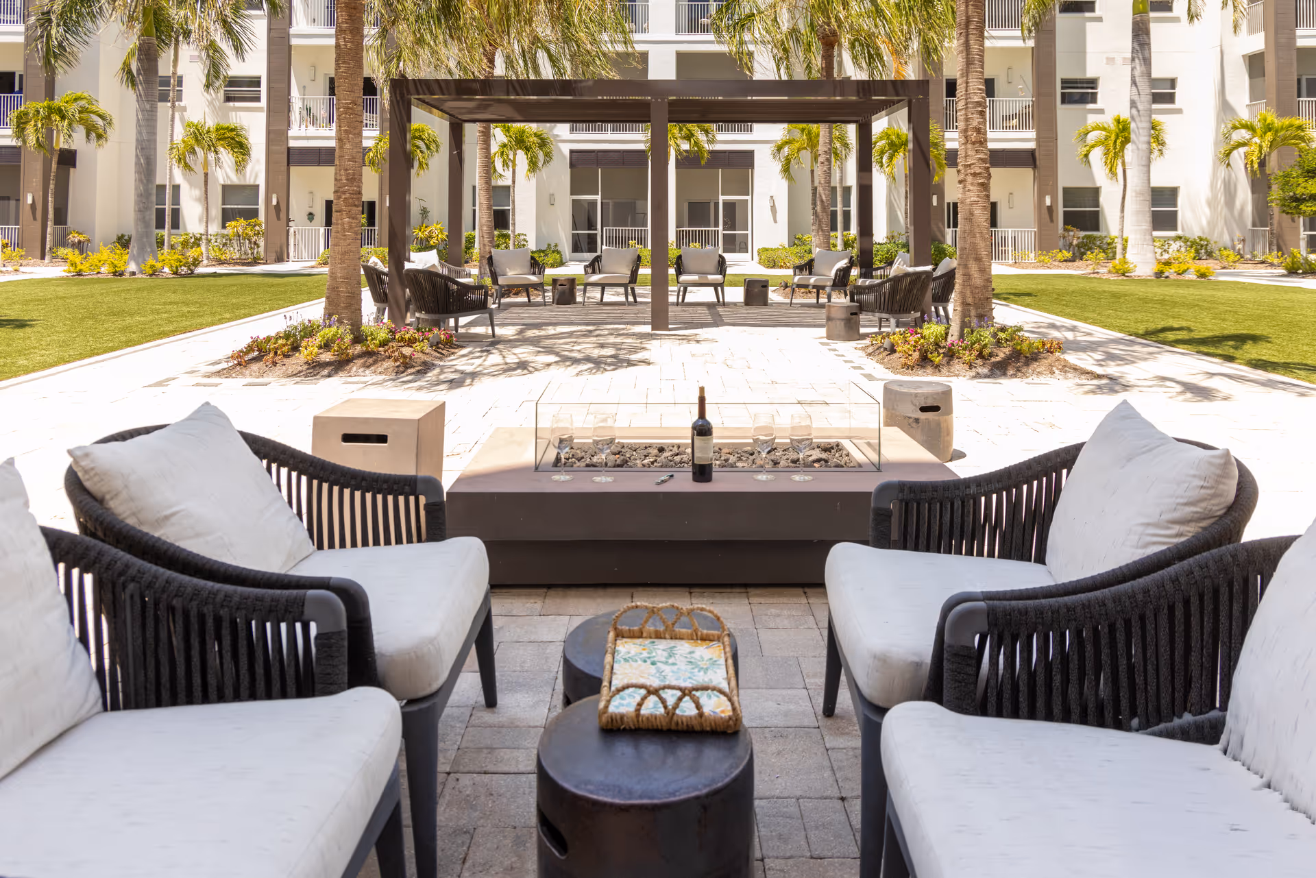 Outdoor seating area at Amavida at Lakes Park featuring cushioned chairs arranged around a rectangular fire pit with a bottle of wine and glasses on it. The area is surrounded by palm trees, green lawns, and a modern building in the background with balconies and large windows.