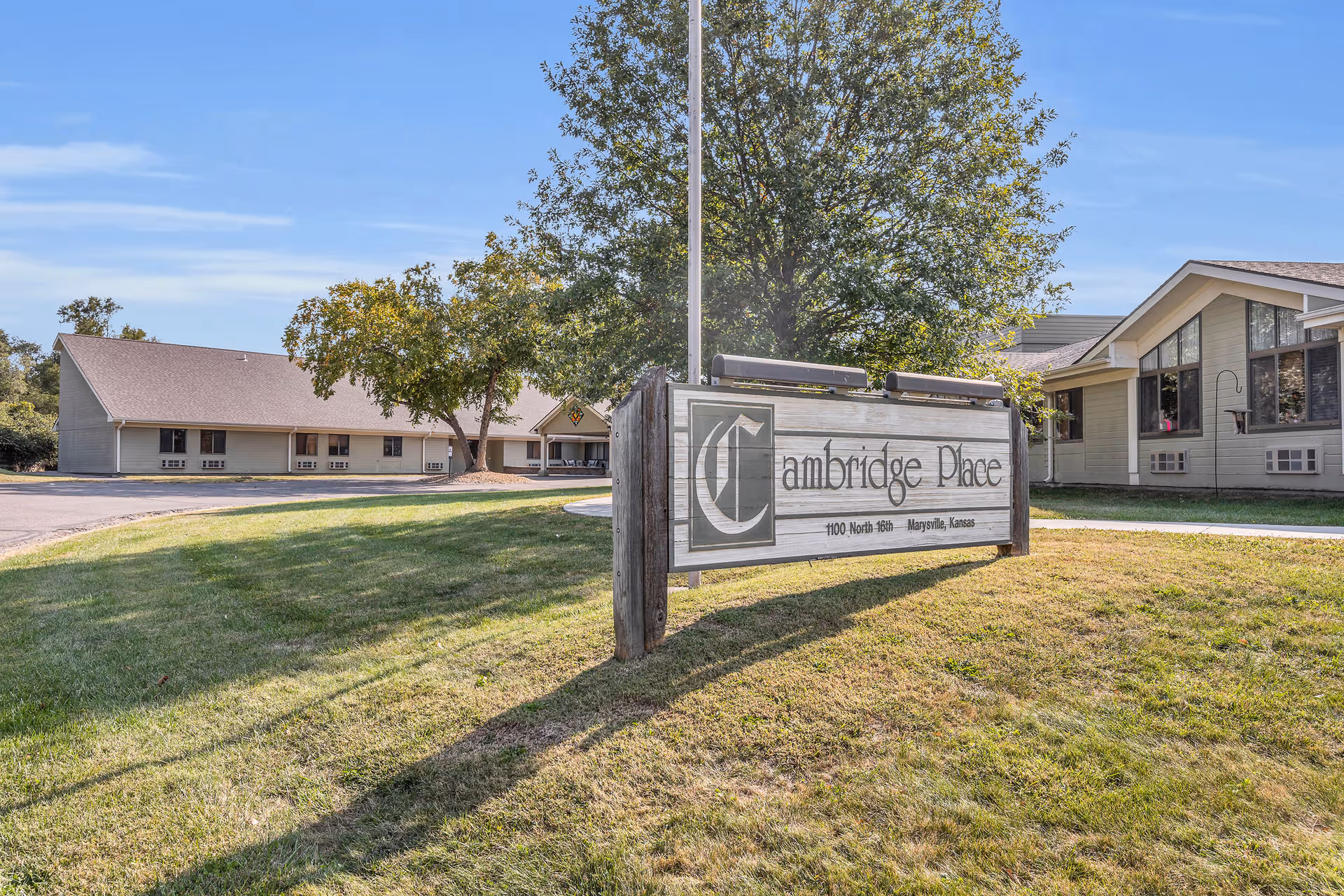 Exterior view of Cambridge Place with a wooden entrance sign on a grassy lawn and the facility buildings behind it.