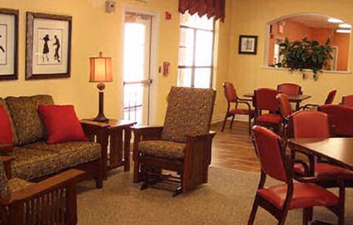 A cozy interior room with a seating area featuring a patterned sofa and armchair with red cushions, a wooden side table with a lamp, and several tables with red chairs in the background. The walls are painted yellow and decorated with framed artwork and a window with a plant visible through an interior pass-through.