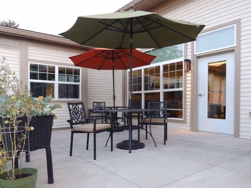 Outdoor patio area with two tables, each shaded by a large umbrella, one green and one red. There are four black metal chairs with beige cushions around the tables. The patio is adjacent to a beige building with large windows and a white door. There are potted plants on the left side of the patio.