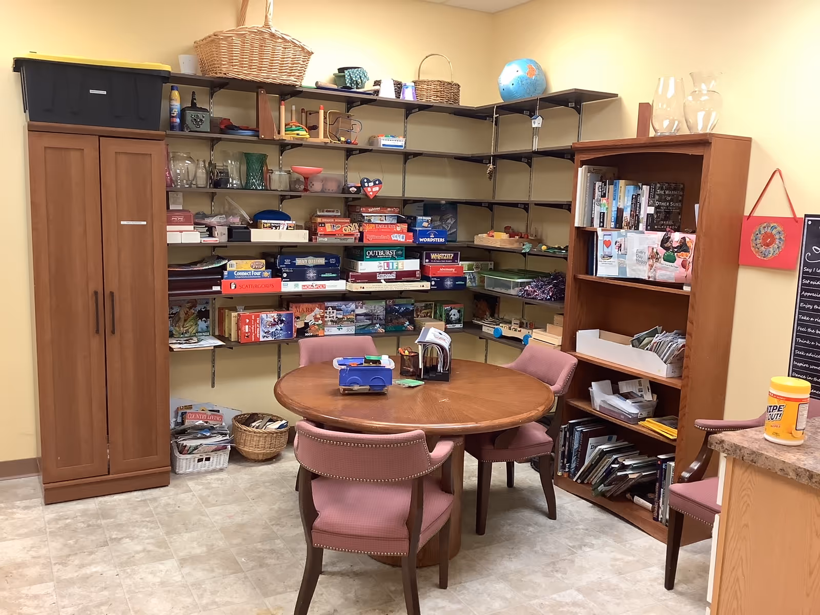 A cozy activity room with a round wooden table surrounded by four pink cushioned chairs. Behind the table, there are shelves filled with board games, puzzles, books, and various small items. A wooden cabinet and a bookshelf are also visible, along with a globe on the top shelf. The room has a tiled floor and light yellow walls.