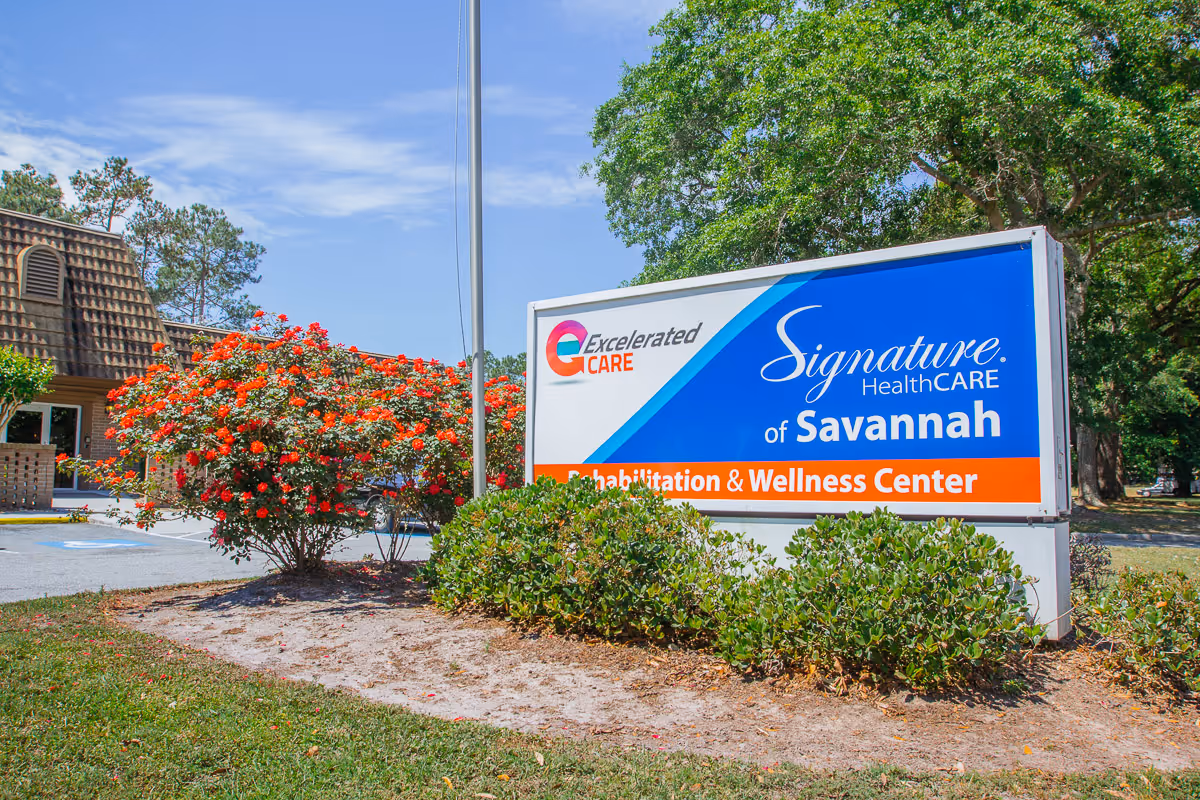 Outdoor front sign for Signature HealthCARE of Savannah next to shrubs and a flowering bush with the facility building in the background.