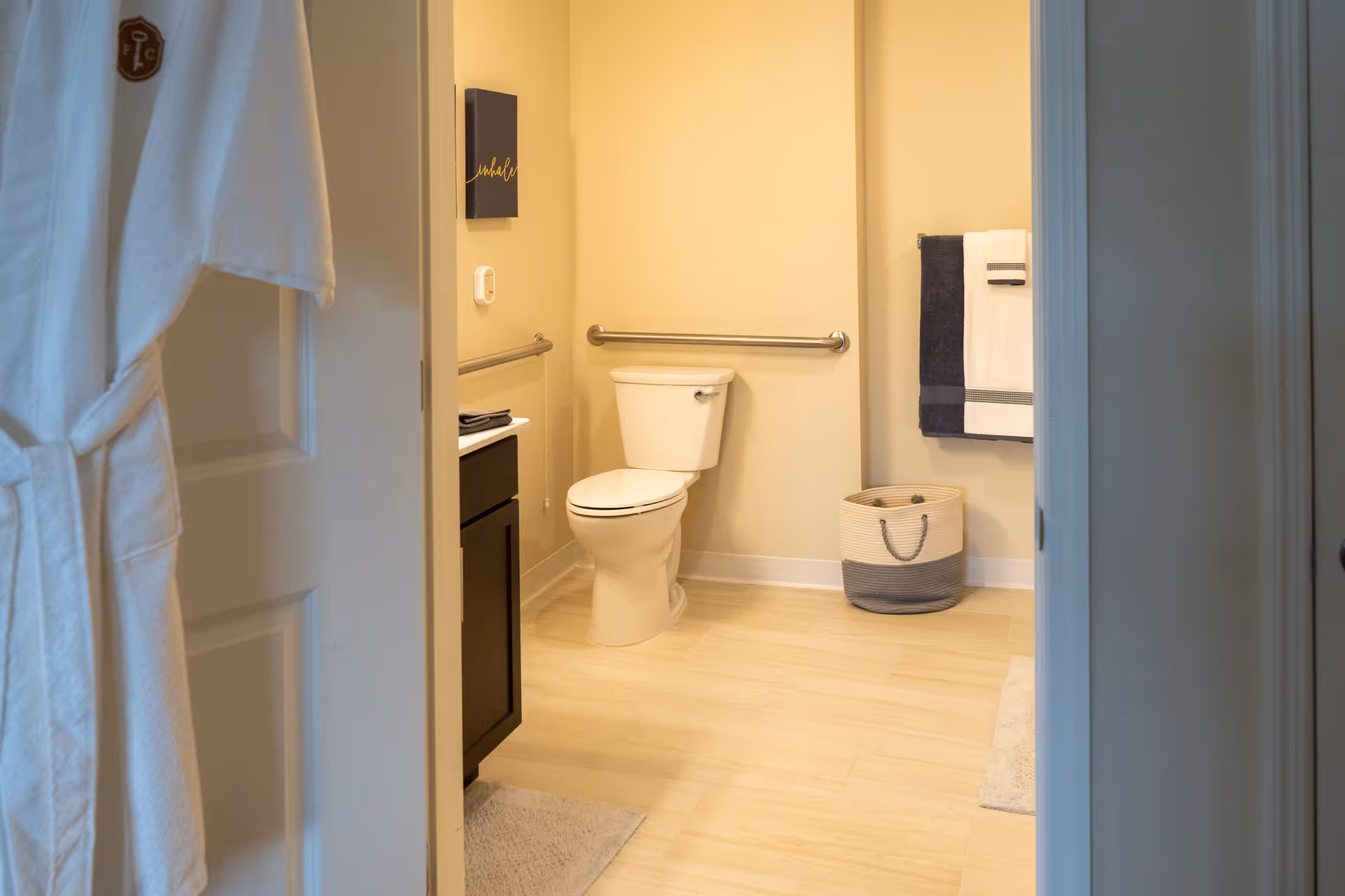View into a bathroom with a white toilet, beige walls, light wood flooring, a dark wood vanity with a white countertop, a towel rack with white and dark blue towels, a woven basket on the floor, and a white bathrobe hanging on the door.