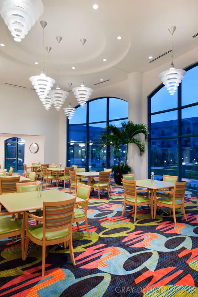 A brightly lit dining room with multiple tables and chairs featuring green cushions and wooden frames. The room has large arched windows showing an outdoor view at dusk. Modern white pendant lights hang from the ceiling, and a colorful patterned carpet covers the floor. There is a large potted plant near a white column.