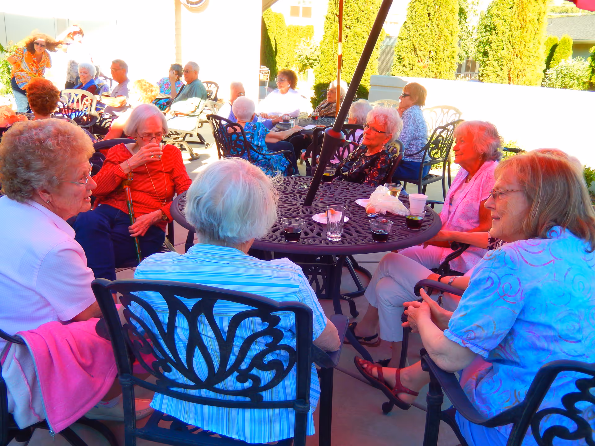 A group of elderly people sitting around outdoor metal tables with drinks, engaging in conversation on a sunny patio with greenery and buildings in the background.
